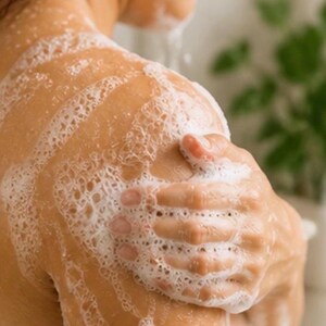 May include: Close-up of a person's shoulder and arm covered in white, foamy soap. The person's hand is gently touching the shoulder, with the soap creating a bubbly texture. The skin appears clean and smooth, suggesting a skincare or bath product.