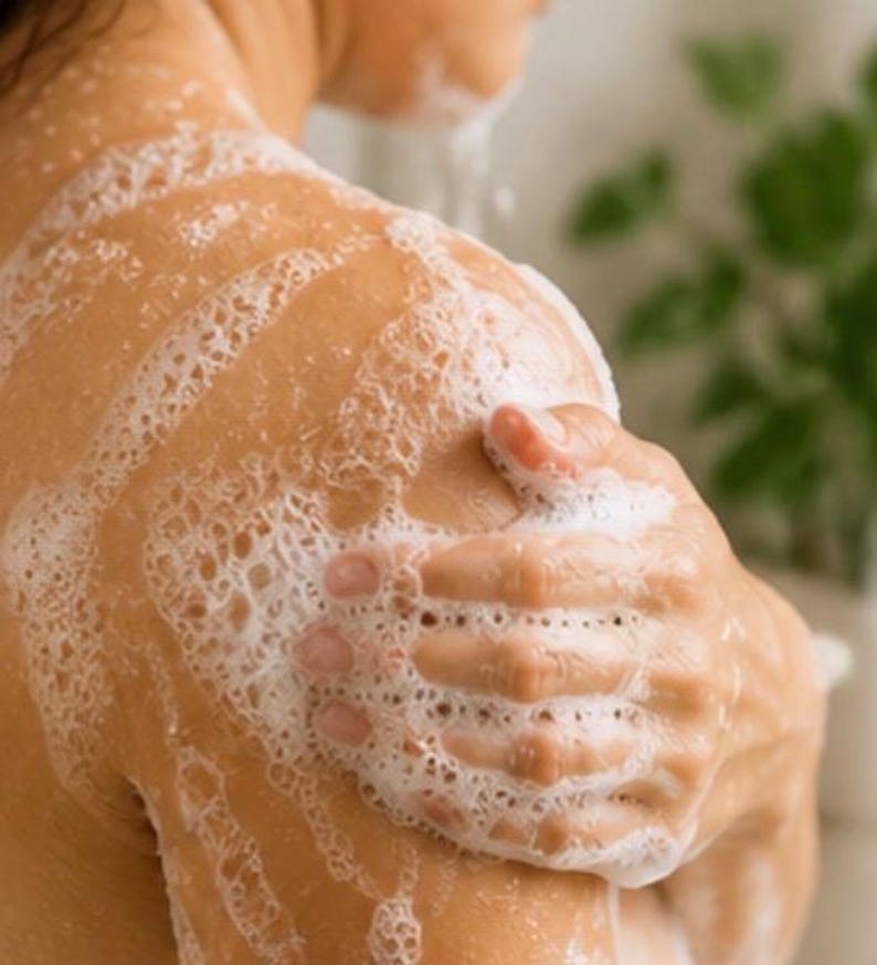 May include: Close-up of a person's shoulder and arm covered in white, foamy soap. The person's hand is gently touching the shoulder, with the soap creating a bubbly texture. The skin appears clean and smooth, suggesting a skincare or bath product.