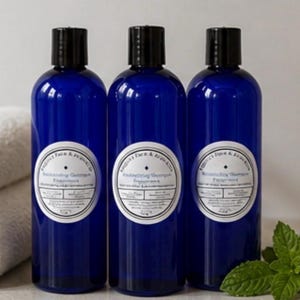 Puede incluir: Tres botellas azul cobalto con tapas negras, cada una con una etiqueta blanca. Las botellas están en fila, con toallas blancas a la izquierda y una ramita de menta a la derecha. Las etiquetas dicen "Moisturizing Shampoo".