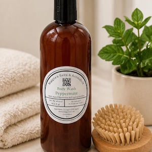 May include: A brown bottle of peppermint body wash with a black cap, labeled "Matgis's Herb & Aromatics." A wooden brush and a small potted plant are next to the bottle. The label states "With Pure Essential & Fragrance Oils."