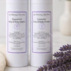 May include: Two white cylindrical bottles of baby and body powder with purple labels. One is labeled "Unscented," the other "Lavender." The labels include text and a small logo. A sprig of lavender and a white towel are in the foreground.