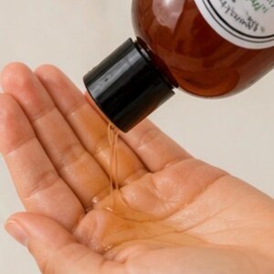 May include: A brown bottle with a black cap is pouring a clear liquid into a hand. The bottle has a white label with black text. The liquid is a light amber color.