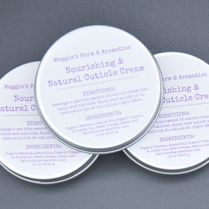 May include: Three round, silver-colored tins of cuticle cream are arranged on a gray surface. Each tin has a white label with purple text that reads "Nourishing & Natural Cuticle Cream." The labels also include directions and ingredients.