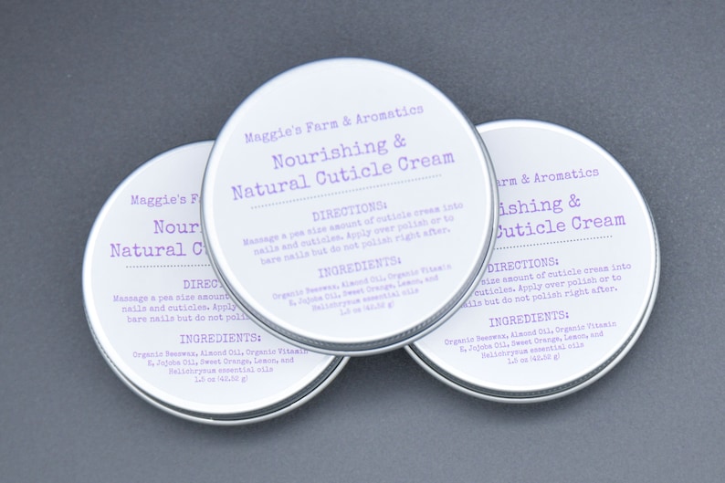 May include: Three round, silver-colored tins of cuticle cream are arranged on a gray surface. Each tin has a white label with purple text that reads "Nourishing & Natural Cuticle Cream." The labels also include directions and ingredients.