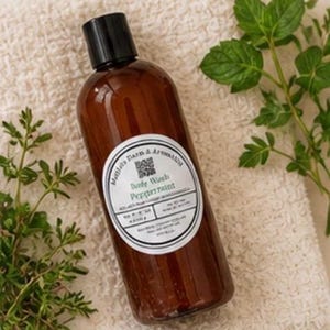 May include: A brown bottle of body wash with a black cap and a white label that reads "Body Wash Peppermint." The bottle is on a white towel with green sprigs of mint and other herbs.