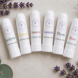May include: Six white aromatherapy inhalers labeled Eucalyptus, Lemon, Cinnamon, Lavender, Thyme, and Rose. Each inhaler has a circular logo and the words "aromatherapy inhaler" printed below the scent name. The image includes sprigs of lavender.