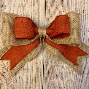 May include: A large burlap and orange ribbon bow. The bow is tied with a knot in the center and has two long tails.