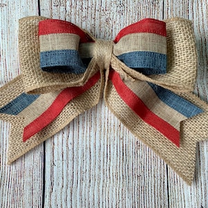 May include: A large burlap bow with red, white, and blue ribbon accents. The bow is tied in a knot and has two long tails.