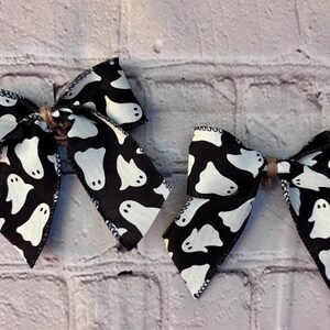May include: Two Halloween-themed hair bows. Each bow is made of black fabric with a white ghost pattern. The bows are tied with twine in the centre. The bows are set against a white brick background.
