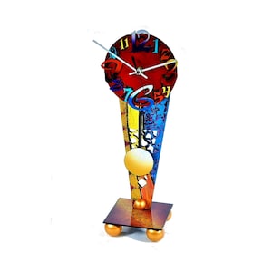 May include: A colourful abstract clock with a red face and multi-coloured numbers. The clock is mounted on a gold and blue metal stand with a round ball at the top. The stand has a square base with four gold balls.