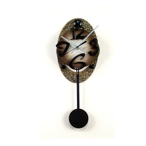 May include: A brown and black pendulum clock with a unique design. The clock face is oval shaped and features black numbers that are offset from the centre. The clock has a black pendulum and a black metal frame.
