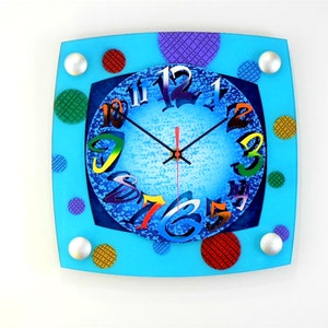 Puede incluir: Un reloj cuadrado azul con números coloridos y lunares. La esfera del reloj tiene un fondo azul texturizado con puntos blancos.