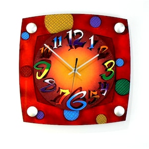 May include: A red square clock with colorful numbers and a white background. The clock has a white face with black hands and a red frame. The clock is decorated with colorful polka dots.