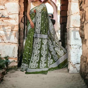 May include: A woman wearing a green and white patterned saree with silver accents. The saree is draped over her shoulders and around her waist. She is standing in a doorway with stone walls on either side.