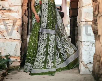 May include: A woman wearing a green and white patterned saree with silver accents. The saree is draped over her shoulders and around her waist. She is standing in a doorway with stone walls on either side.