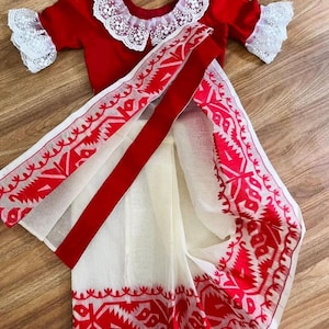 Puede incluir: Una blusa roja tradicional con ribete de encaje blanco y un sari blanco y rojo a juego. El sari presenta intrincados patrones rojos a lo largo de los bordes y una amplia franja roja. El conjunto se exhibe sobre una superficie de madera.