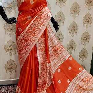 May include: A bright orange silk saree with a white intricate design. The saree has a traditional Indian pattern with geometric shapes and floral motifs.