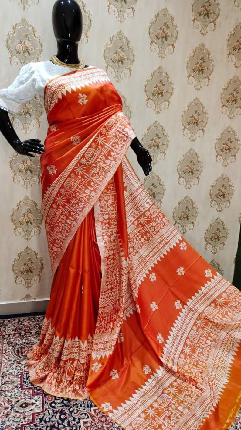 May include: A bright orange silk saree with a white intricate design. The saree has a traditional Indian pattern with geometric shapes and floral motifs.