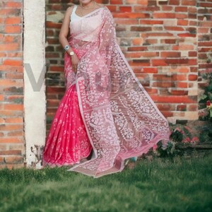 May include: A pink and white saree with intricate embroidery. The saree is draped over a person standing in front of a brick wall.