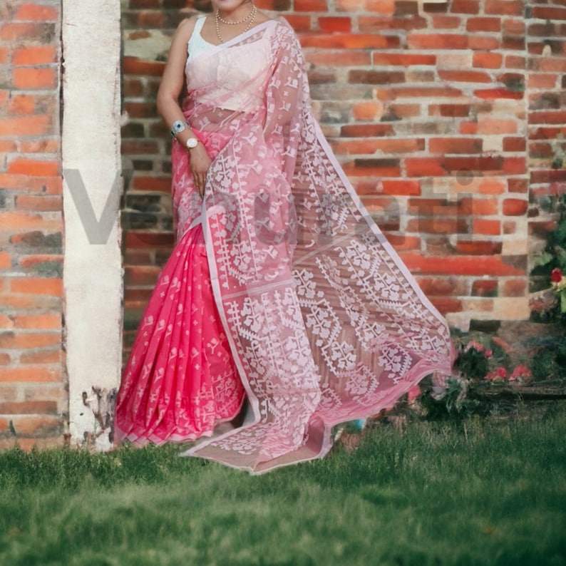 May include: A pink and white saree with intricate embroidery. The saree is draped over a person standing in front of a brick wall.