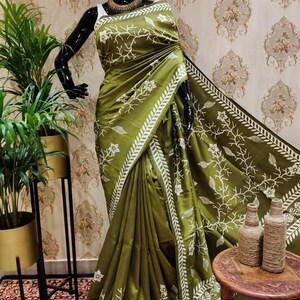 May include: A green silk saree with white floral embroidery. The saree has a white border with a leaf design. The saree is draped on a mannequin.