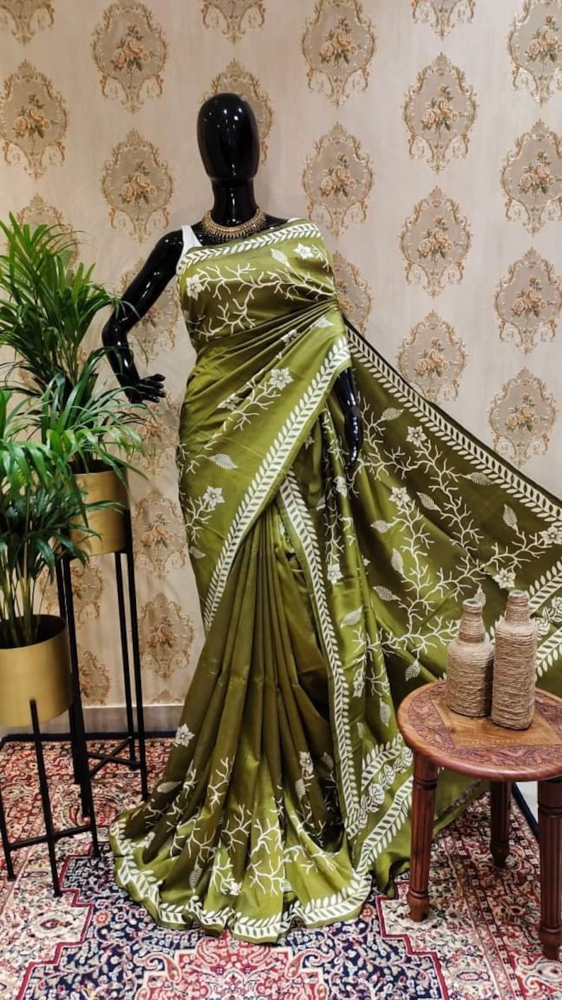 May include: A green silk saree with white floral embroidery. The saree has a white border with a leaf design. The saree is draped on a mannequin.