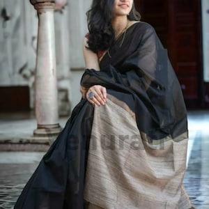 May include: A woman wearing a black saree with a gold border. The saree is draped over her shoulders and waist, and she is sitting on a tiled floor.