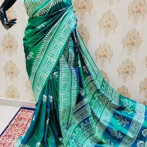May include: A green and blue silk saree with a white floral pattern. The saree is draped on a mannequin and is displayed on a patterned rug.