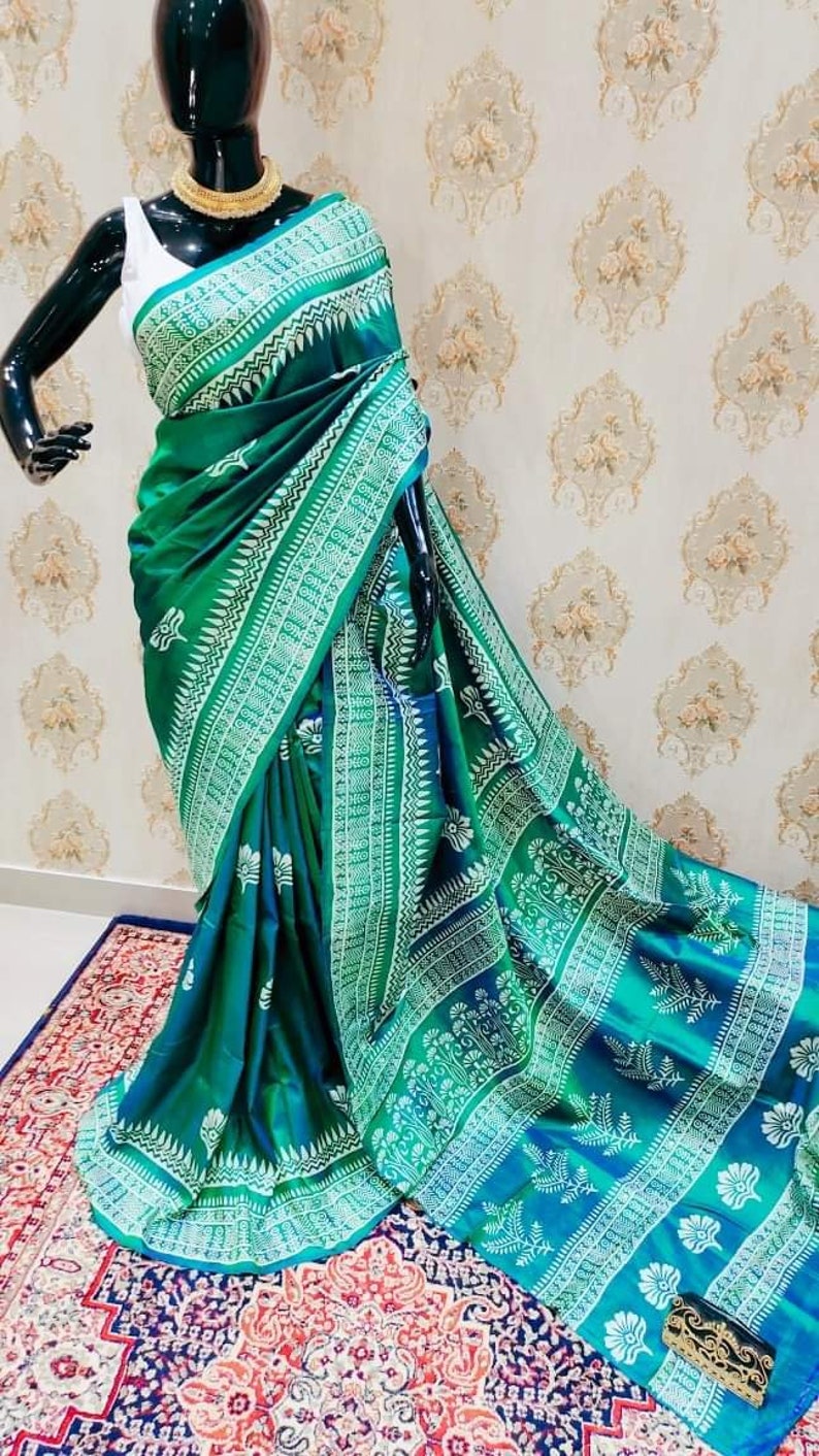 May include: A green and blue silk saree with a white floral pattern. The saree is draped on a mannequin and is displayed on a patterned rug.