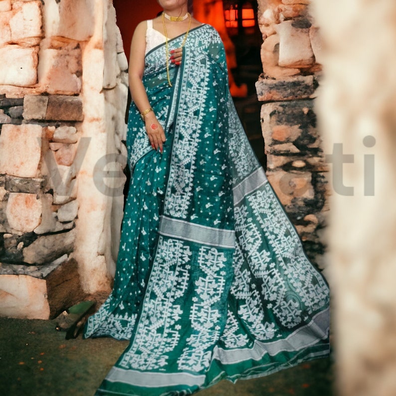May include: A green and white patterned saree with a silver border. The saree is draped around a person standing in front of a stone wall.