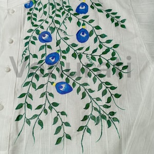 May include: A white shirt with a hand-painted design of blue flowers and green vines. The design is on the front of the shirt, near the collar.