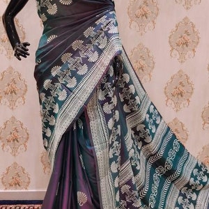 May include: A dark purple and teal silk saree with a white geometric pattern. The saree is draped on a mannequin and has a gold necklace.