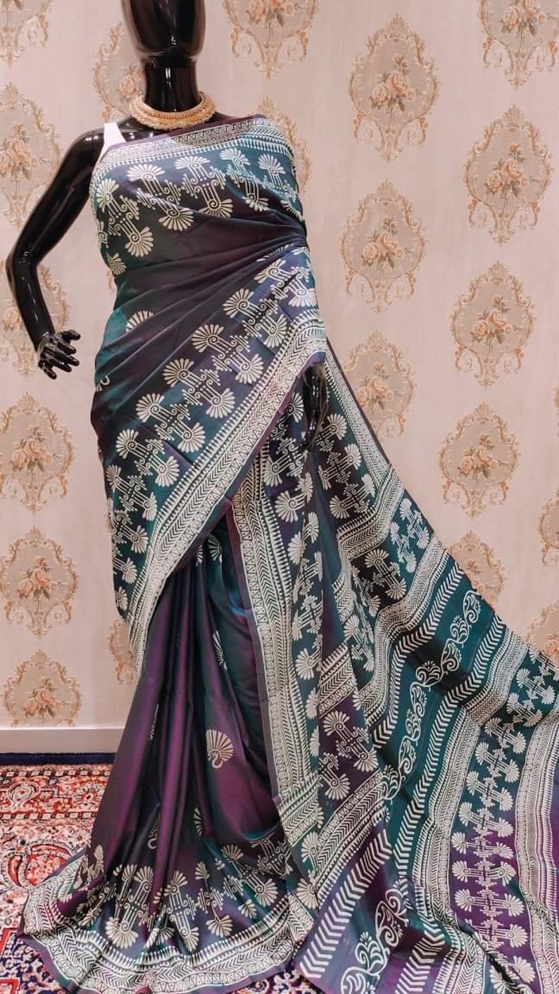 May include: A dark purple and teal silk saree with a white geometric pattern. The saree is draped on a mannequin and has a gold necklace.