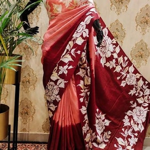 May include: A maroon and peach silk saree with a white floral print. The saree is draped on a mannequin and has a gradient effect from peach to maroon.