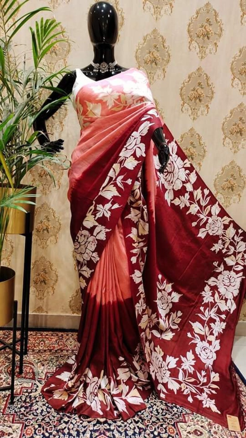 May include: A maroon and peach silk saree with a white floral print. The saree is draped on a mannequin and has a gradient effect from peach to maroon.