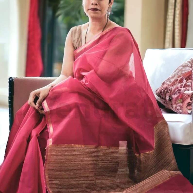 May include: A woman wearing a pink saree with gold trim. The saree is draped over a white chair with a floral patterned pillow.