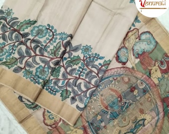 Pode incluir: Um sari de seda cor creme com uma borda dourada e desenhos intrincados pintados à mão. Os desenhos apresentam flores azuis, folhas verdes e um motivo central de uma divindade em uma moldura circular.