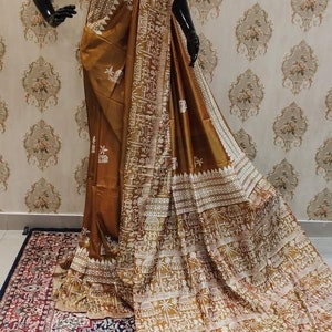 May include: A brown silk saree with a white embroidered design featuring geometric patterns and stylized figures. The saree has a gold border and is draped on a mannequin.