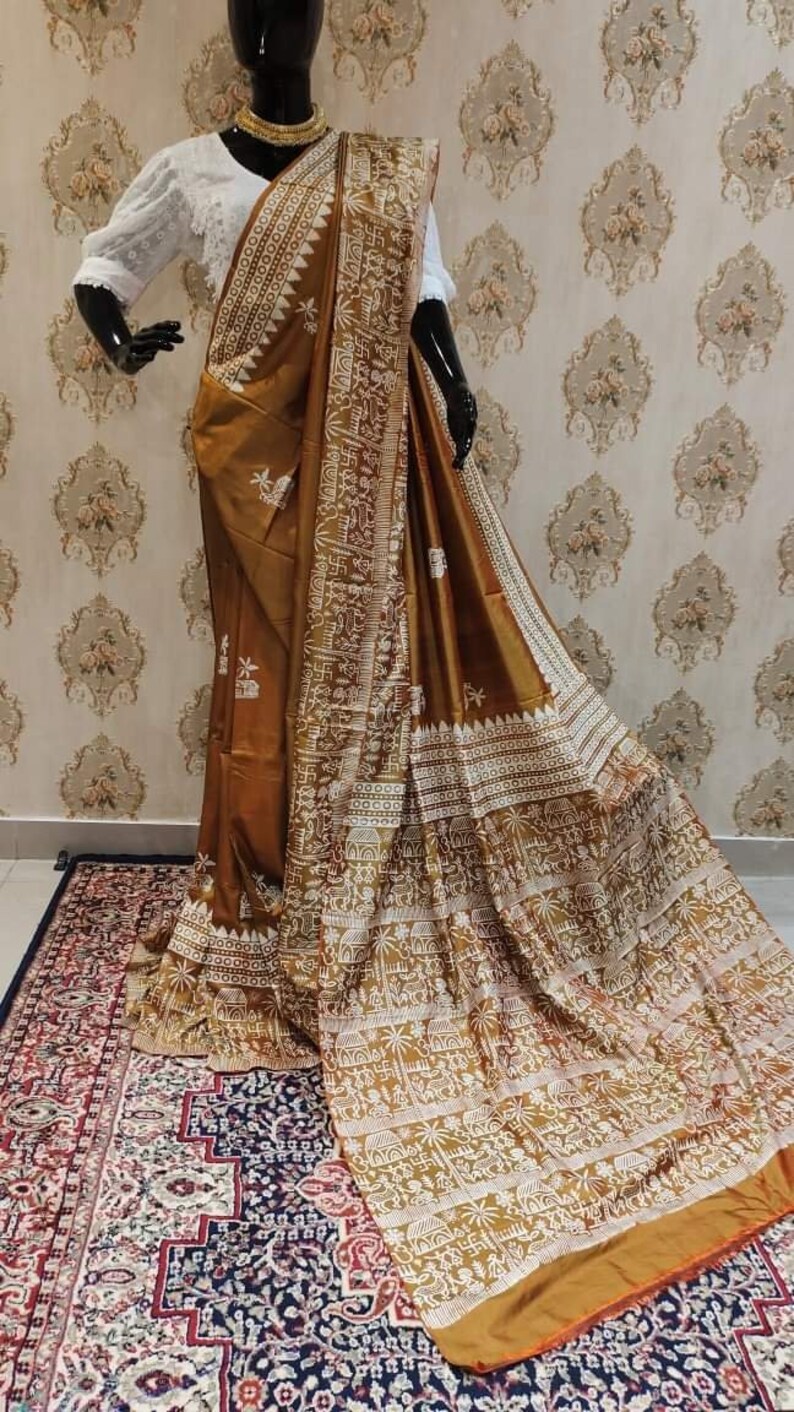 May include: A brown silk saree with a white embroidered design featuring geometric patterns and stylized figures. The saree has a gold border and is draped on a mannequin.
