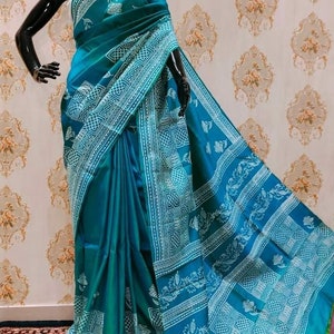 May include: A teal blue and white silk saree with a traditional block print design. The saree has a gradient effect, with the teal blue color fading to a lighter shade of blue at the bottom. The saree is draped on a mannequin.