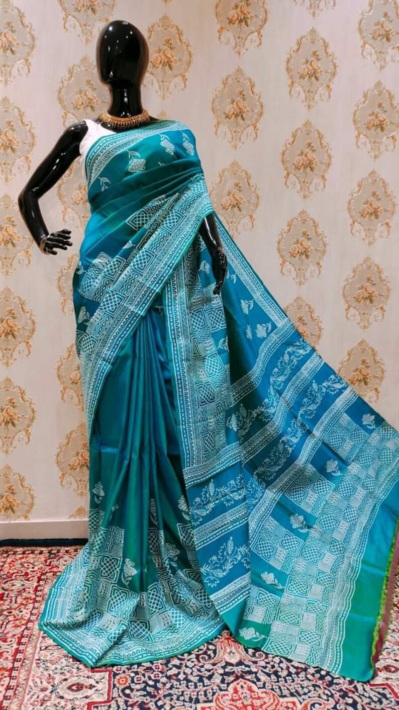 May include: A teal blue and white silk saree with a traditional block print design. The saree has a gradient effect, with the teal blue color fading to a lighter shade of blue at the bottom. The saree is draped on a mannequin.