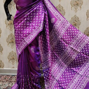 May include: A purple silk saree with a white floral print. The saree has a gradient effect, with the color fading from a darker purple to a lighter purple. The saree is draped over a mannequin.