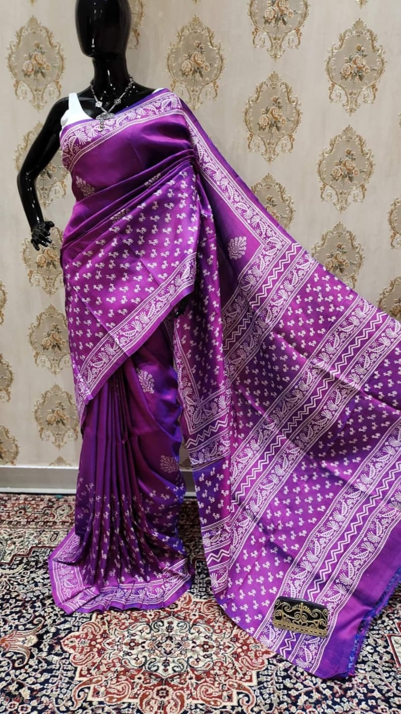 May include: A purple silk saree with a white floral print. The saree has a gradient effect, with the color fading from a darker purple to a lighter purple. The saree is draped over a mannequin.