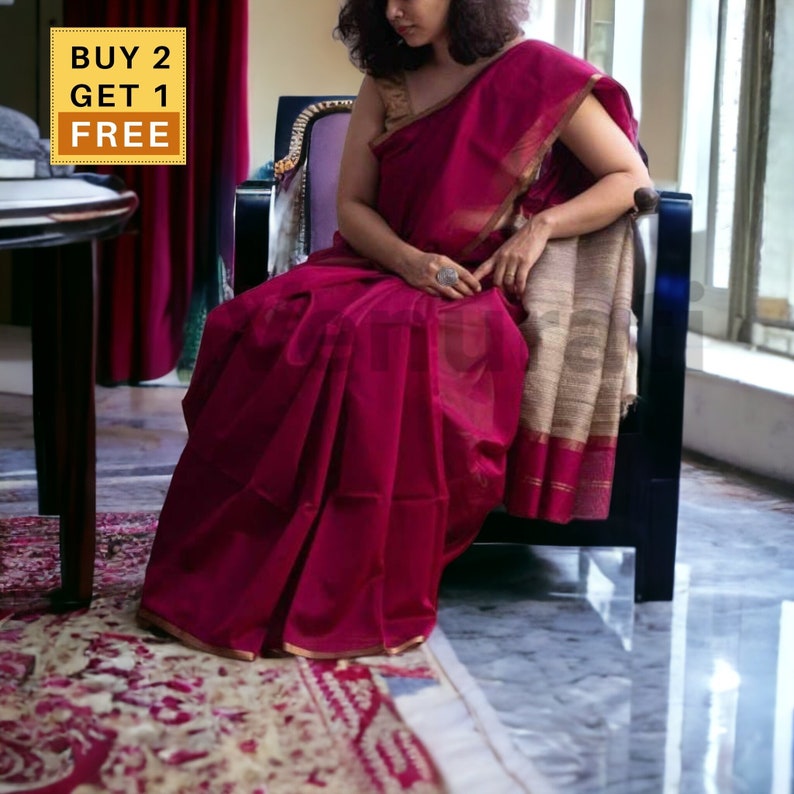 May include: A woman wearing a deep burgundy saree with a gold border and a beige and gold patterned pallu. The saree is draped over a chair and the woman is sitting on the chair. The saree has a red and gold border. The woman is wearing a gold ring on her finger. The image has the text "BUY 2 GET 1 FREE" in a yellow box.
