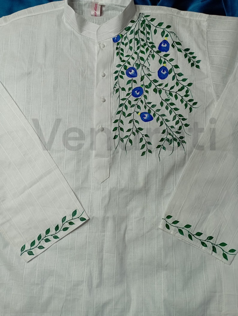 May include: White long-sleeved shirt with green vines and blue flowers painted on the front. The shirt has a mandarin collar and button closure. The sleeves have green vines painted on the cuffs.