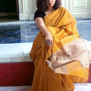 May include: A mustard yellow saree with a cream and gold border. The saree is draped around a person and has a subtle sheen.