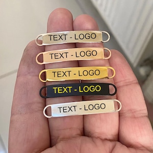 Könnte beinhalten: Fünf Metallanhänger in Silber, Roségold, Gold, Schwarz und Silber. Jeder Anhänger hat eine rechteckige Form mit abgerundeten Enden und einer kleinen Schlaufe. Der Text "TEXT - LOGO" ist auf jedem Anhänger aufgedruckt.