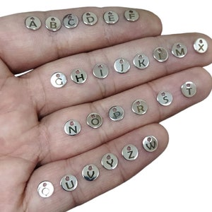 Puede incluir: Un conjunto de abalorios de letras del alfabeto de metal plateado. Los abalorios son redondos y tienen un agujero en la parte superior para sujetarlos a joyas u otras manualidades. Las letras son de la A a la Z y la X.