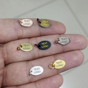 May include: A collection of oval-shaped metal charms in various colors, including gold, silver, and black. Each charm has a small loop for attachment and the text "TEXT LOGO" printed on its surface. The charms are displayed against a neutral background.