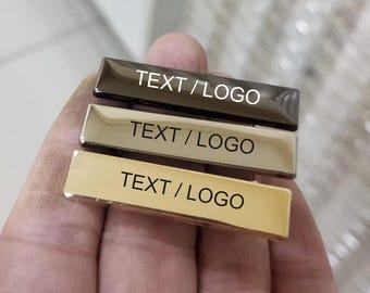 Puede incluir: Tres insignias rectangulares en marrón, beige y dorado, cada una con el texto "TEXT / LOGO" en blanco. Las insignias se sostienen en una mano, mostrando su acabado metálico pulido. Ideal para la personalización.